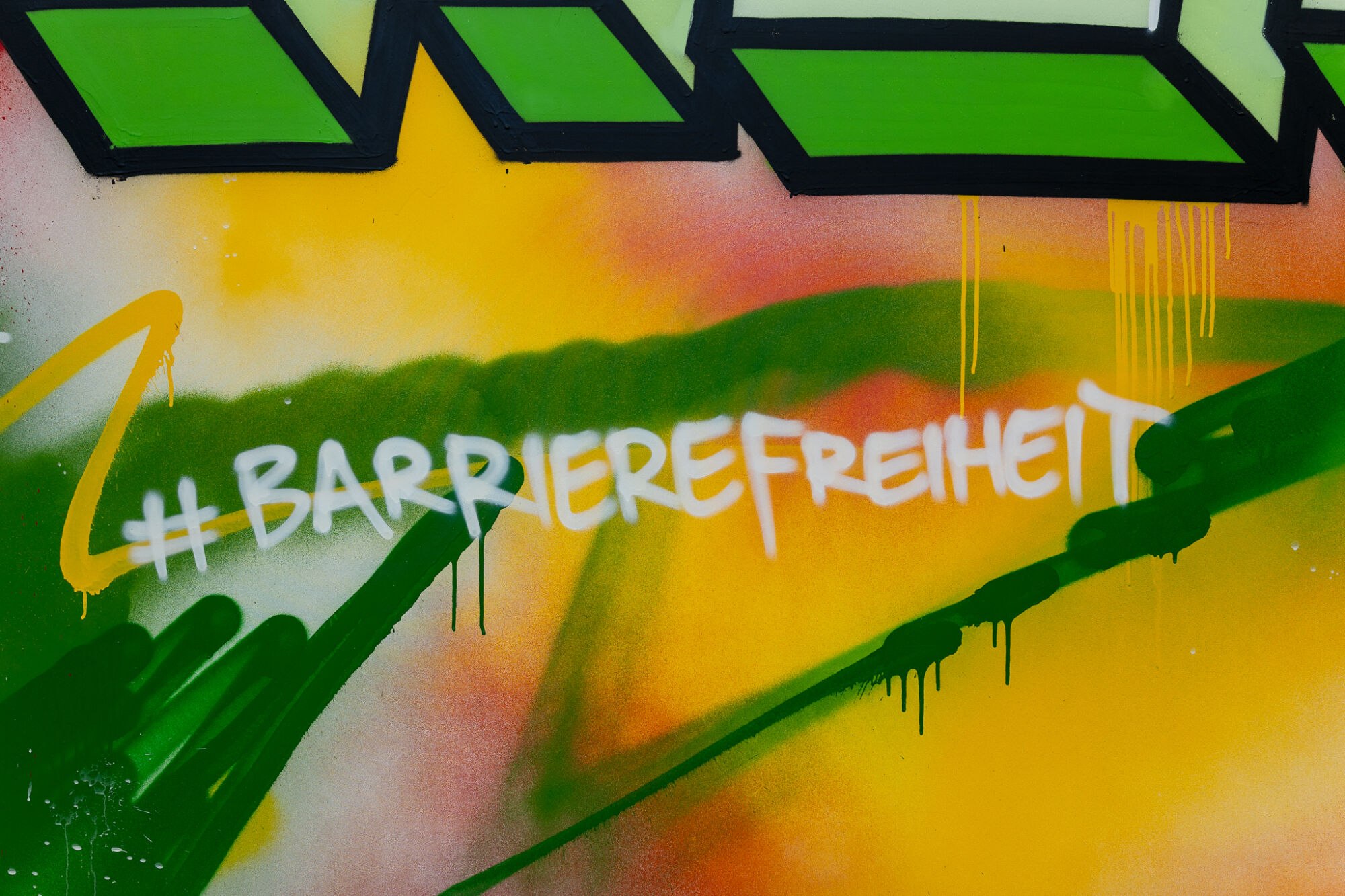 Weißer Schriftzug „#Barrierefreiheit“ auf einem Graffito in Grün, Gelb und Orange.